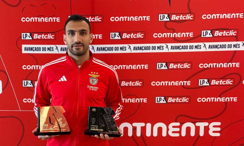 An S.L. Benfica football player standing in front of a red sponsor backdrop, holding two monthly performance awards.