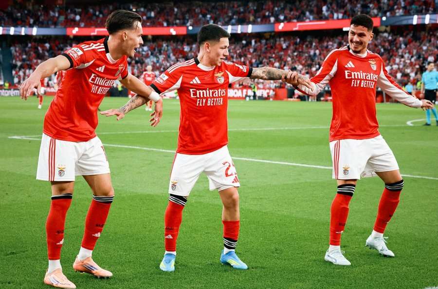 Three S.L. Benfica football players in red home kits smiling and celebrating together on the pitch in front of a crowded stadium.